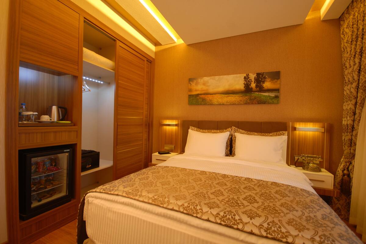 A hotel room with a large bed, wooden wardrobe, a small refrigerator, and a painting on the wall.