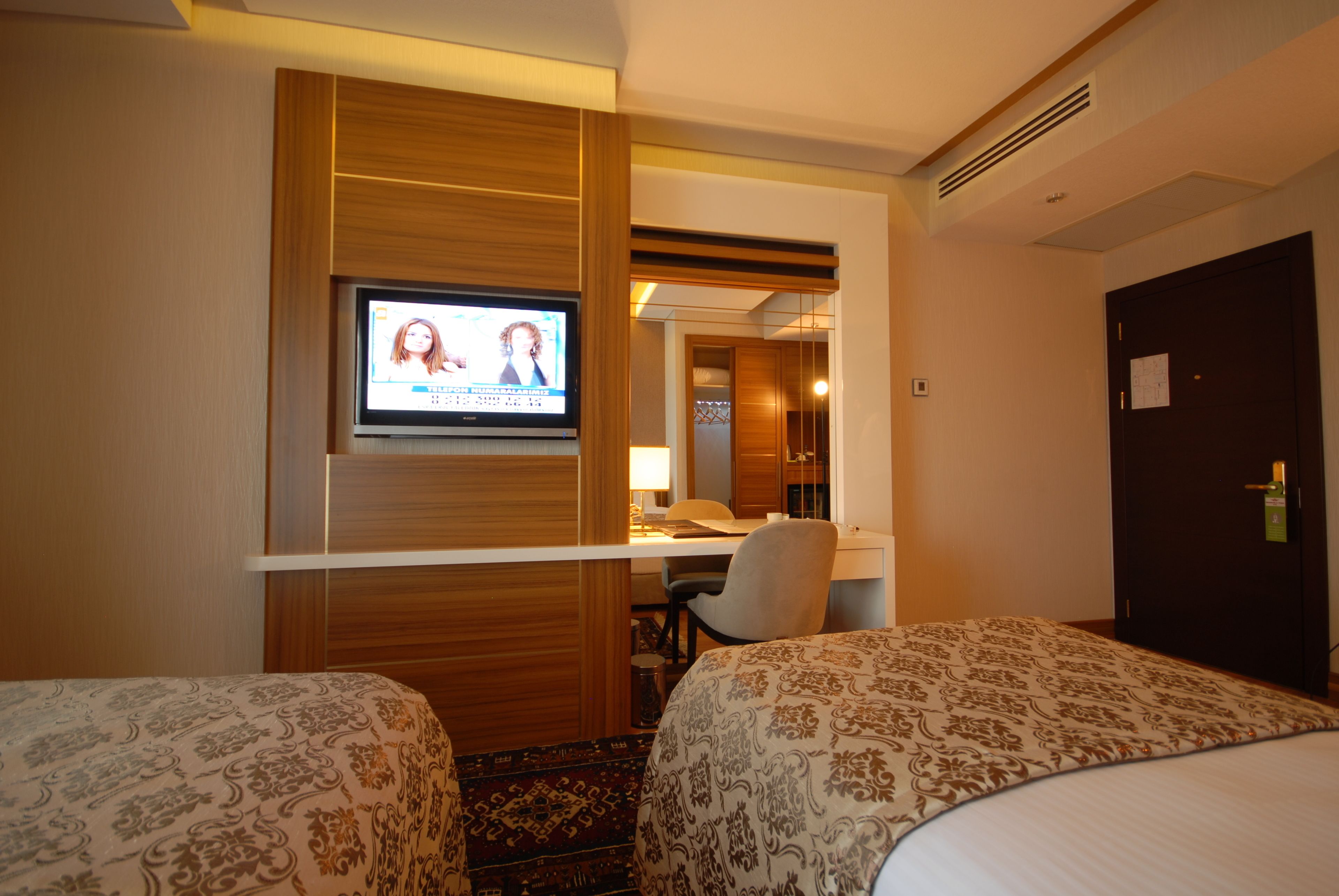 Habitación de hotel con dos camas, un escritorio con silla, televisor montado en la pared y una puerta con cerradura.