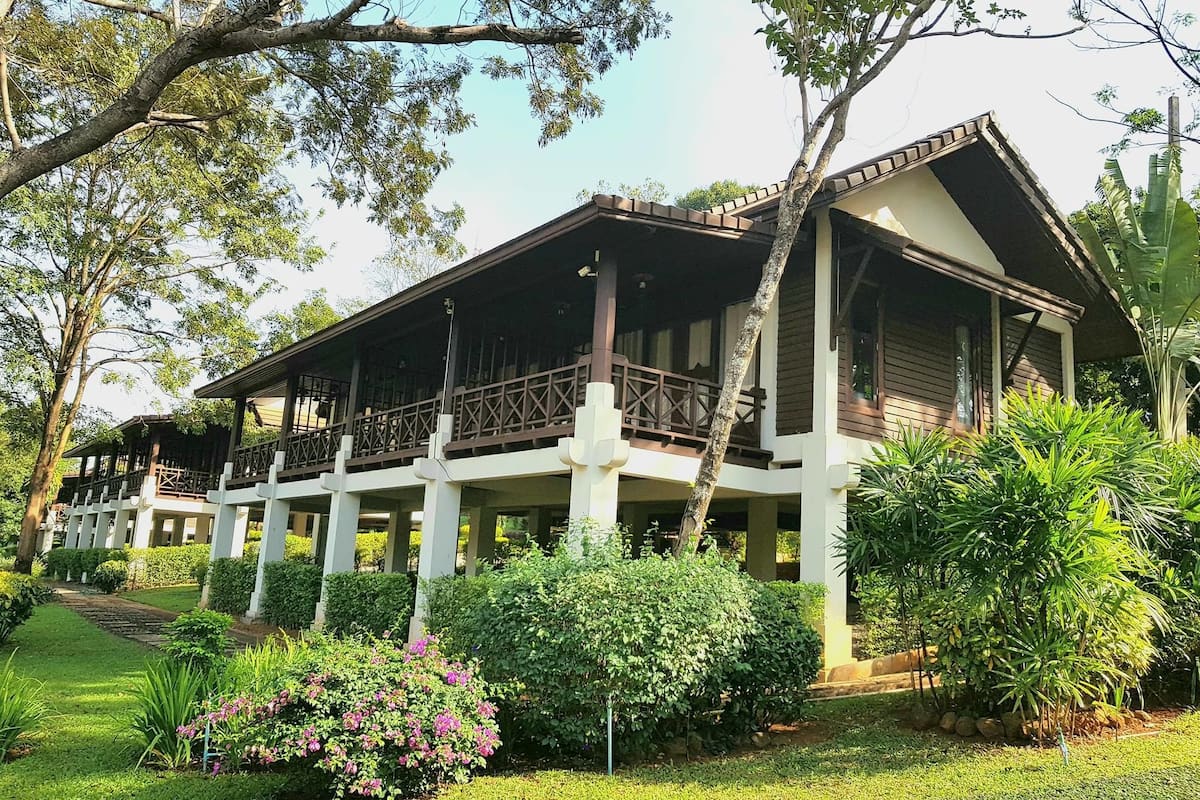 Um edifício de dois andares com varanda, cercado por vegetação exuberante e um jardim bem cuidado.
