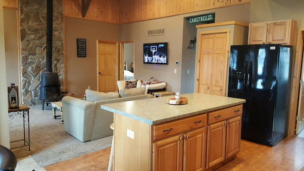 A cozy living room with a stone fireplace, a large TV, and a kitchen area with wooden cabinets and a black refrigerator.