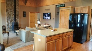 A cozy living room with a stone fireplace, a large TV, and a kitchen area with wooden cabinets and a black refrigerator.