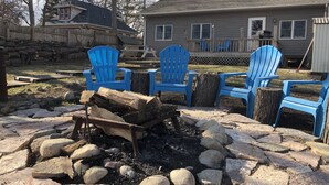 A backyard with a fire pit surrounded by rocks and logs, blue Adirondack chairs, and a wooden deck.