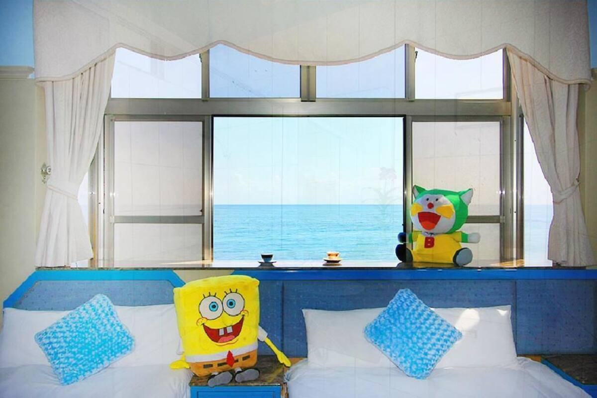 A room with a large window, a bed with blue and white bedding, and a SpongeBob SquarePants plush toy on the bed.