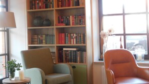 A room with a large wooden bookshelf filled with books, a green armchair, a red armchair, a small table with a candle, and a window with a view of a car outside. - zeitlos -  Hotel & Restaurant am Fuchsbach (Pattensen)