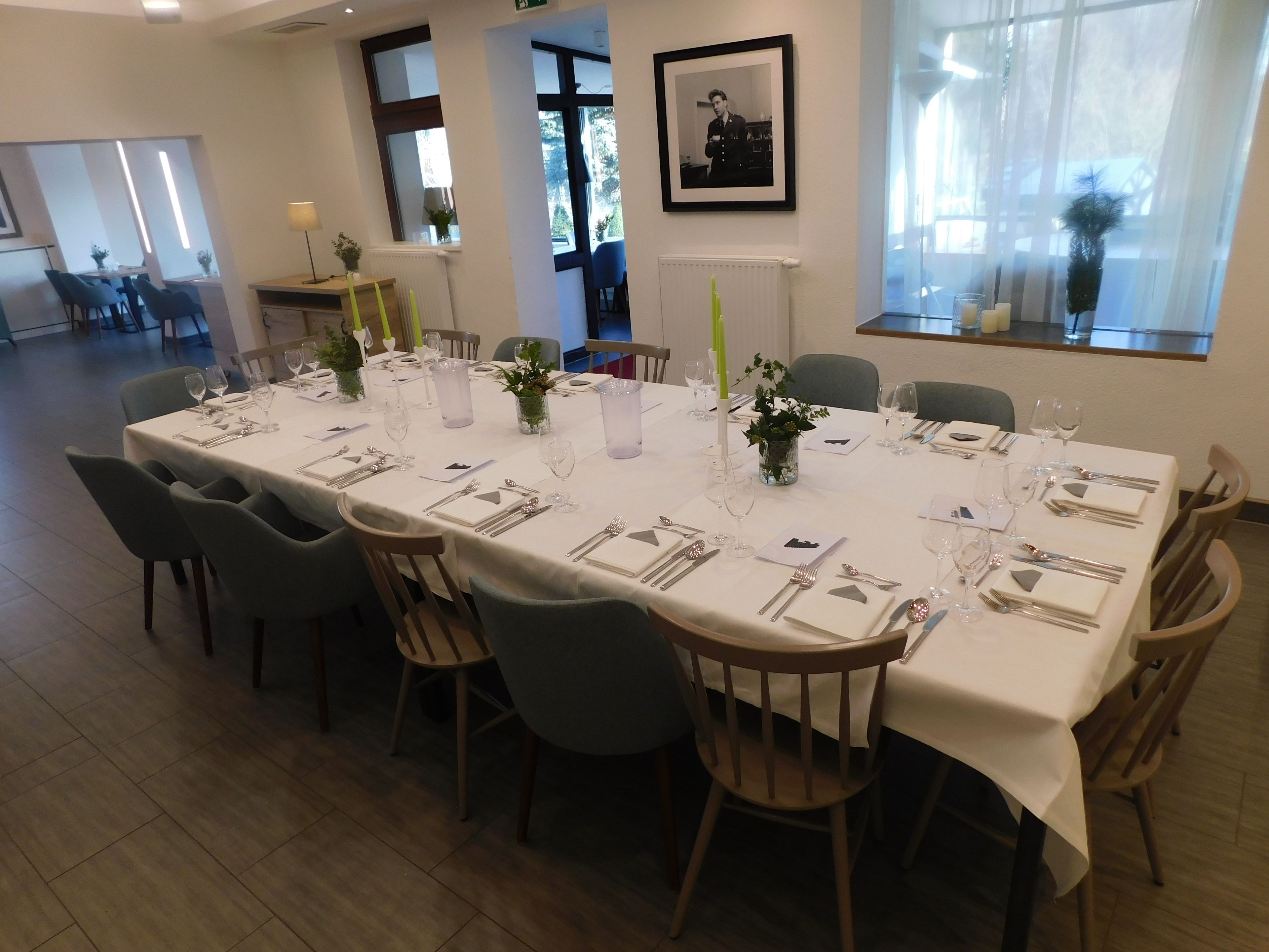 A dining area with a long table set for six, surrounded by chairs, with a window view and a framed picture on the wall.
