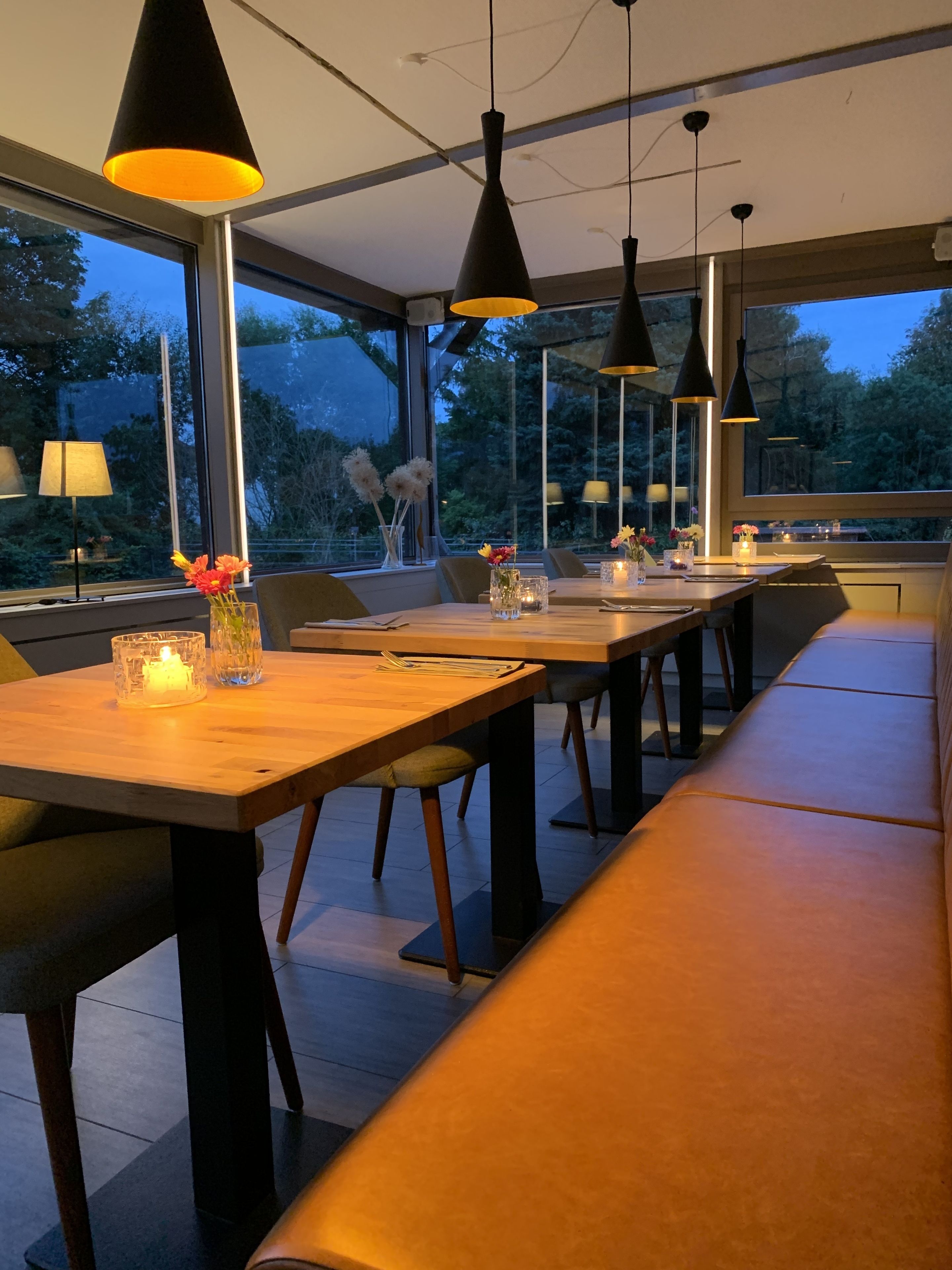 A modern dining area with wooden tables and benches, pendant lighting, and large windows offering a view of the outdoors.