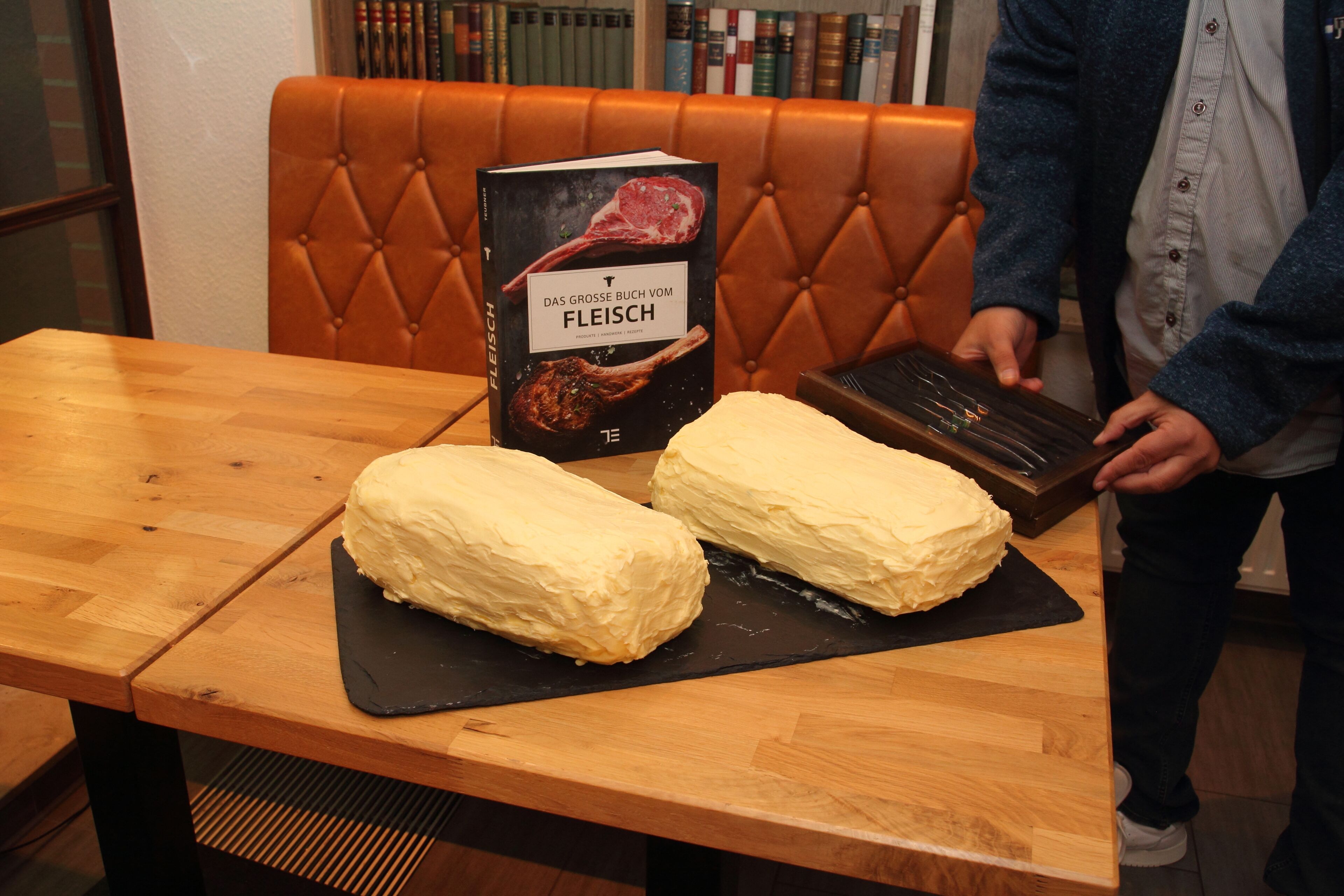 A table with two large, uncut cakes on a black slate, a book titled 'Das Grosse Buch Vom Fleisch' on the table, and a person standing beside the table.