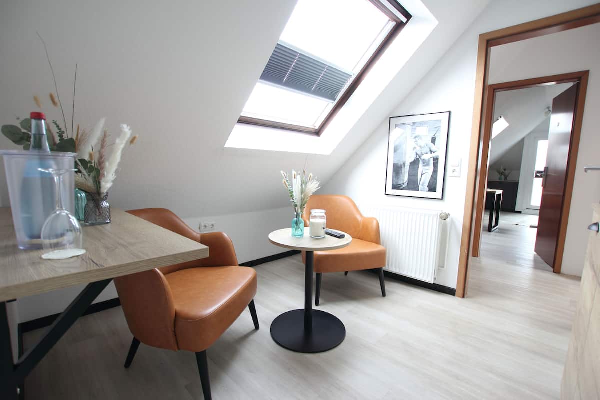 A modern room with a skylight, a dining table with chairs, a small round table with a vase and a candle, and a framed picture on the wall.