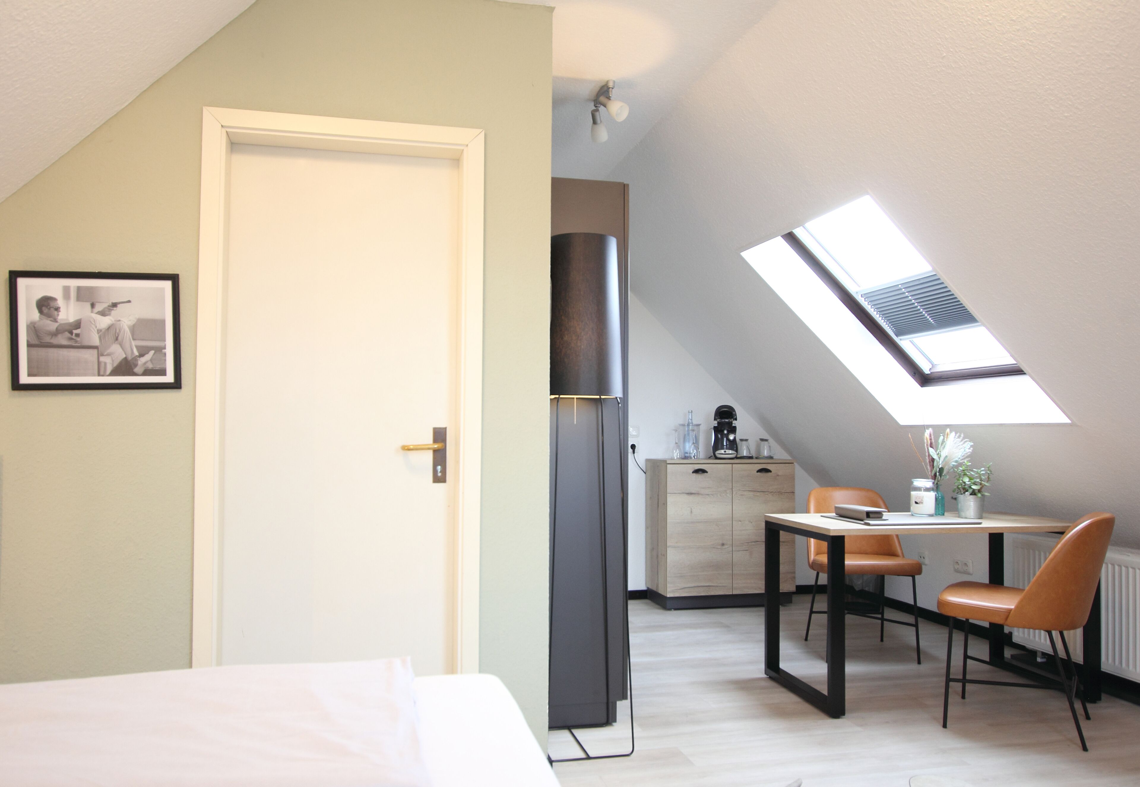 A bedroom with a skylight, a bed, a desk with a chair, and a kitchenette.