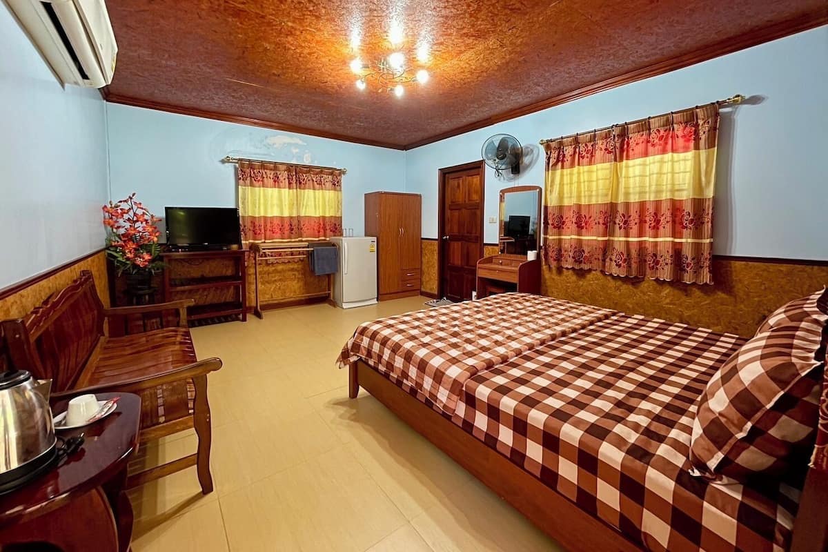 A hotel room with a wooden ceiling, a bed with a checkered bedspread, a desk with a chair, a television, a refrigerator, and a window with curtains.