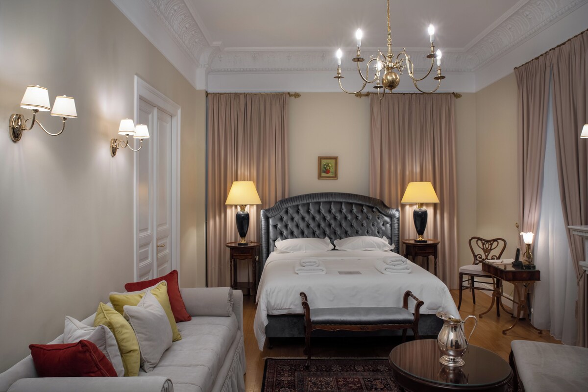 A hotel room with a large bed, two bedside tables with lamps, a sofa, a chair, and a chandelier.
