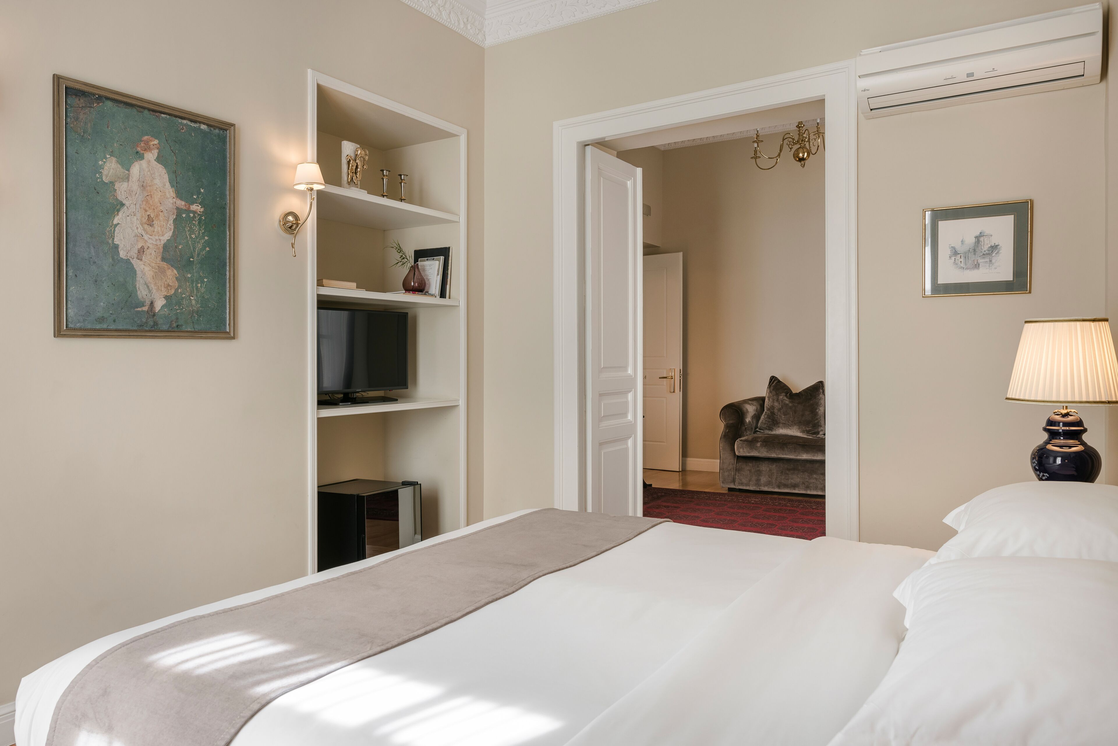A hotel room with a large bed, a television on a shelf, a painting on the wall, and a door leading to another room.