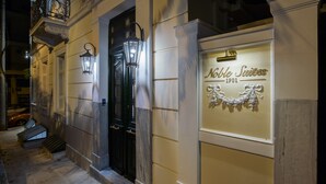 A hotel entrance with a black door, a sign reading "Noble Suites 1901," and a small outdoor seating area. - Noble Suites (Athens)