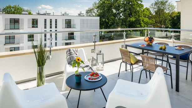 Un espace de restauration extérieur moderne, doté d’une balustrade en verre, de chaises blanches, d’une table dressée avec des fruits et des légumes, et offrant une vue sur les bâtiments environnants.