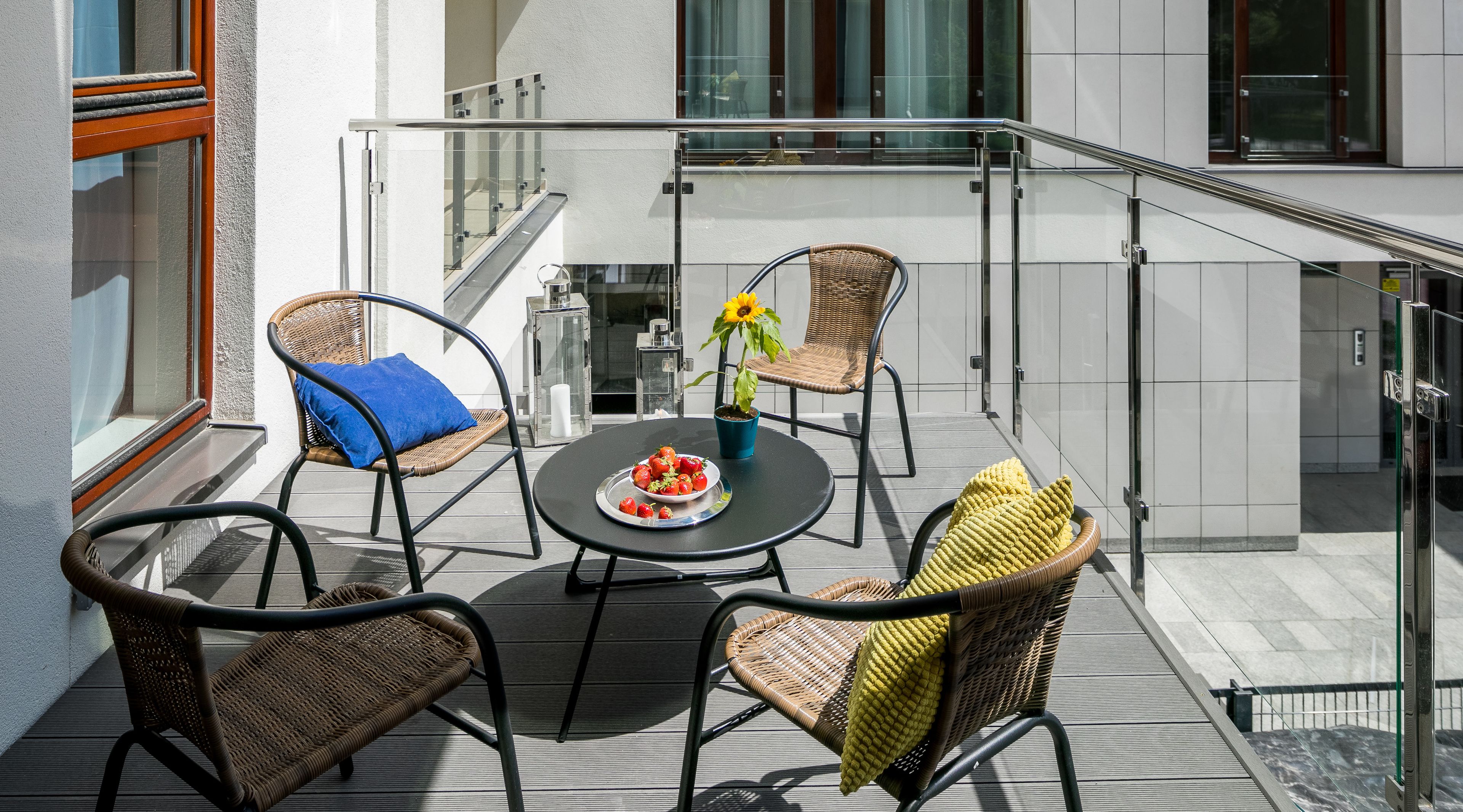 A balcony with wicker chairs, a round table with a plate of food, and a potted sunflower.