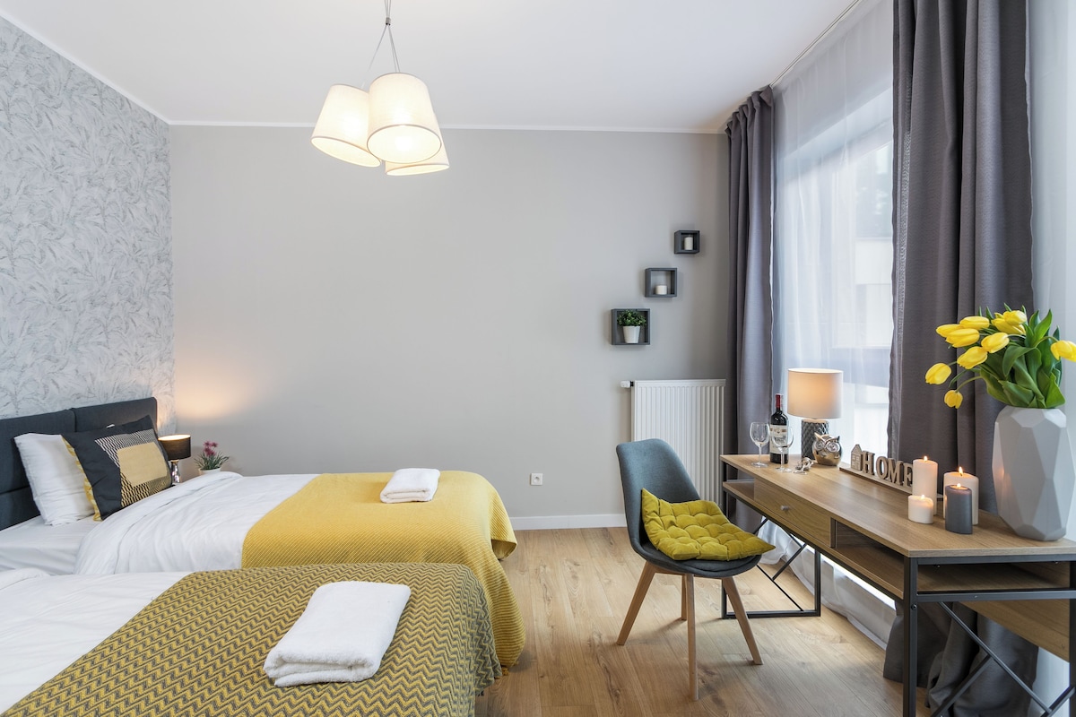 Une chambre d’hôtel avec deux lits, un bureau, une chaise, une fenêtre avec des rideaux et un vase avec des fleurs.