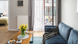 Un salon moderne avec un canapé bleu, une table basse en bois ornée d’un vase de fleurs jaunes, une horloge au mur et une vue sur un balcon extérieur.