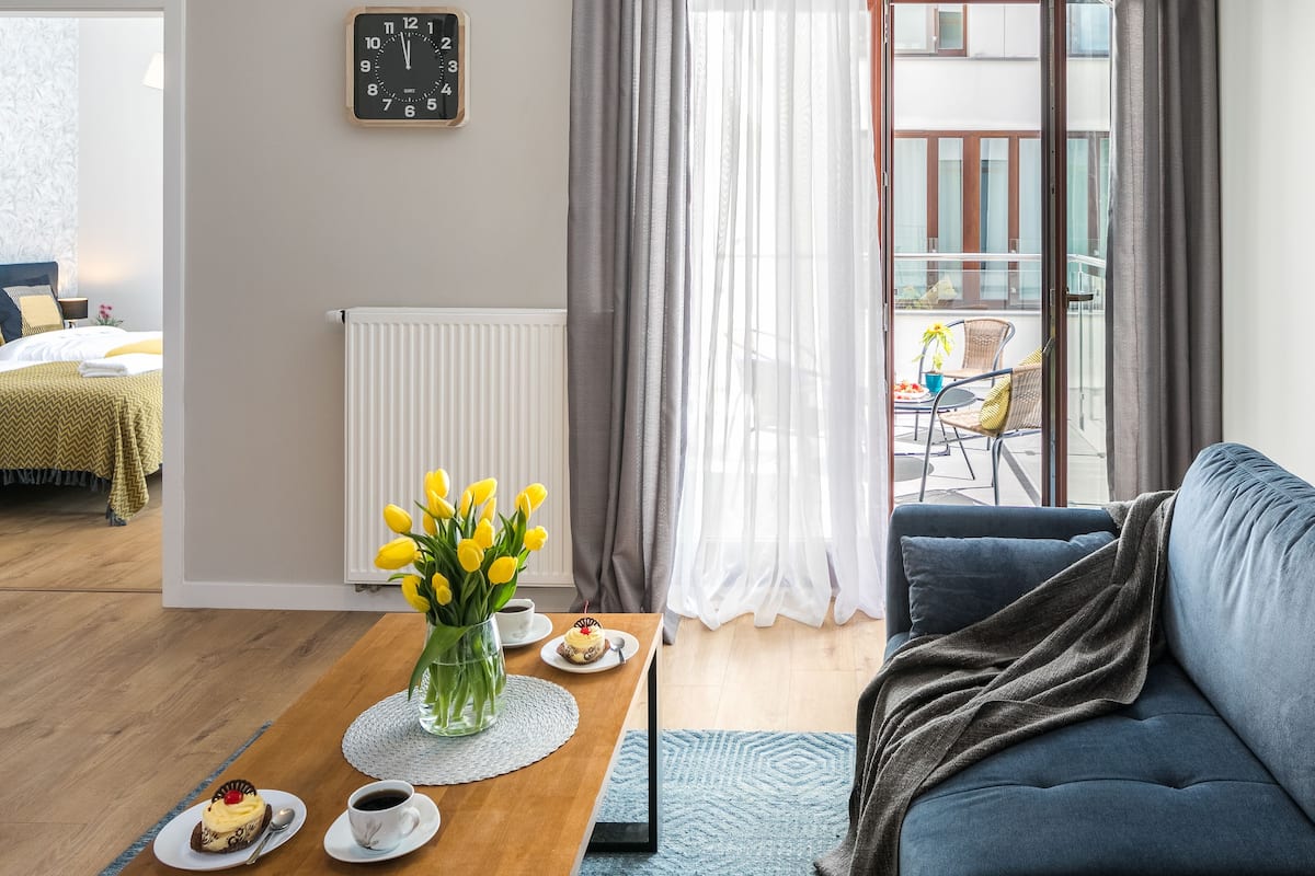 Un salon moderne avec un canapé bleu, une table basse en bois ornée d’un vase de fleurs jaunes, une horloge au mur et une vue sur un balcon extérieur.