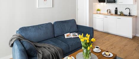 A modern living room with a blue sofa, a coffee table with a vase of yellow tulips, and a small kitchen area with white cabinets and a microwave.