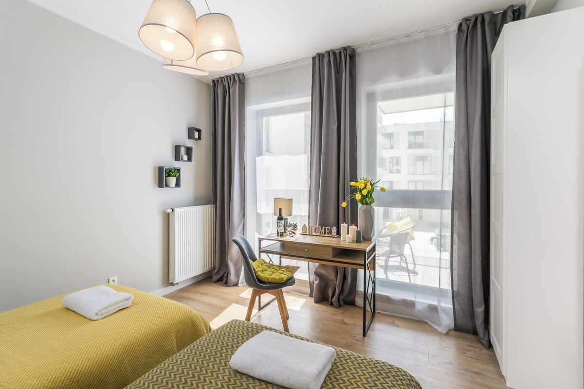 A modern hotel room with two beds, a desk with a chair, a radiator, and a large window with curtains.