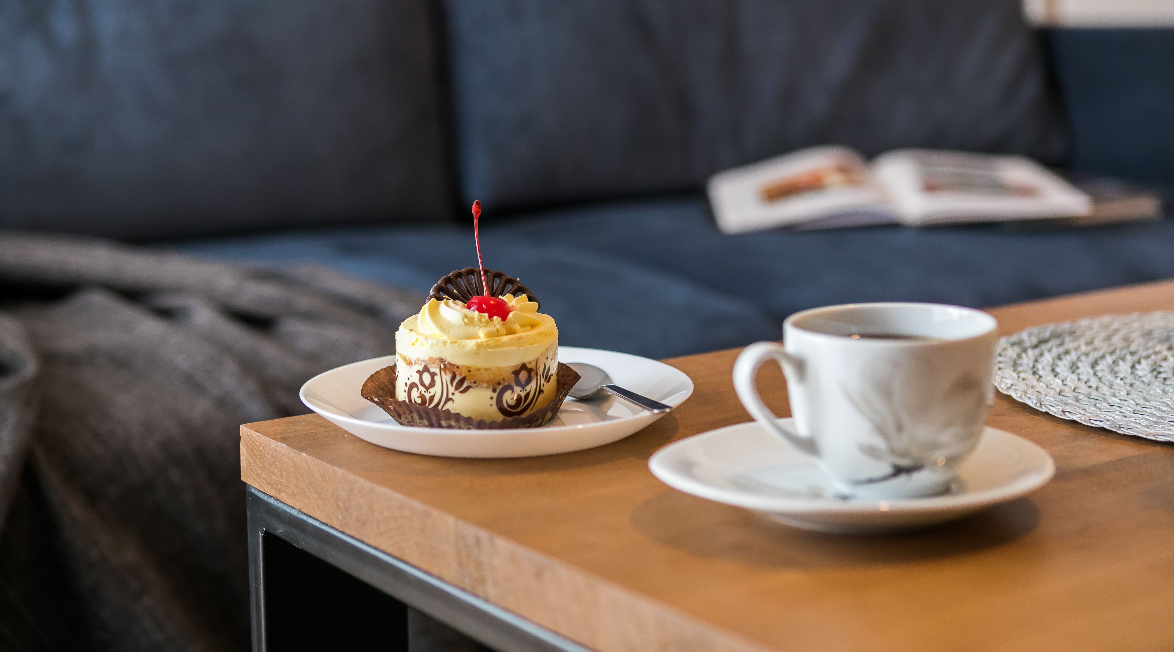 Un dessert présenté sur une assiette, avec une cuillère à côté, une tasse de café sur une soucoupe, et un livre posé sur un canapé à l’arrière-plan.