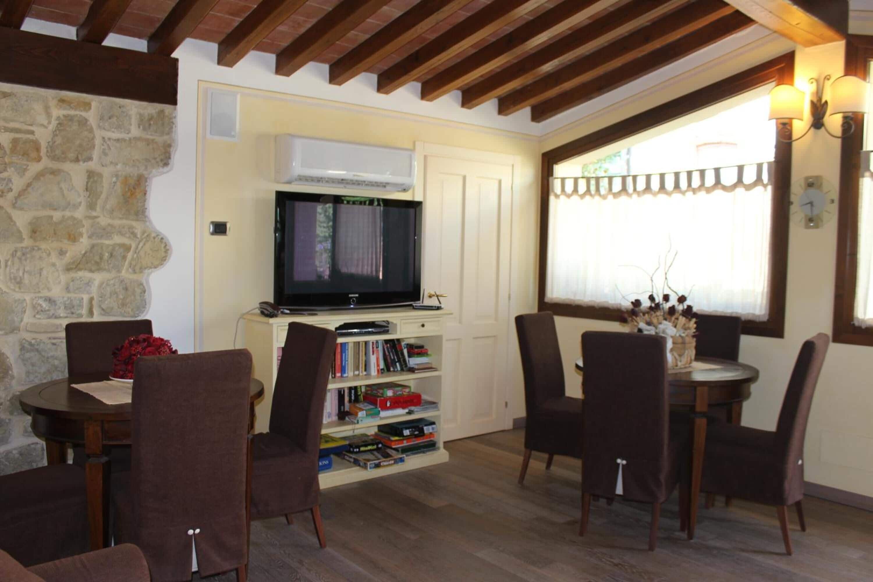 A cozy living room with a stone wall, wooden ceiling beams, a flat-screen TV, a bookshelf, and a dining area with chairs and a table.
