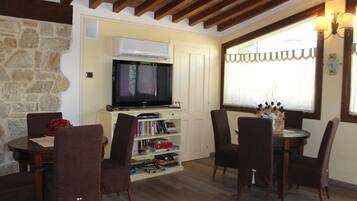 A cozy living room with a stone wall, wooden ceiling beams, a flat-screen TV, a bookshelf, and a dining area with chairs and a table.