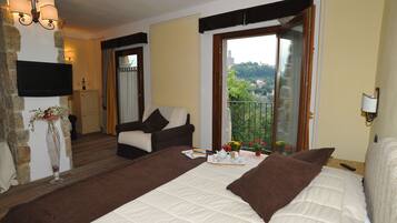 Habitación de hotel con una cama grande, un sofá, una mesita con una bandeja de desayuno, un televisor en una pared de piedra y un balcón con vistas a la vegetación.