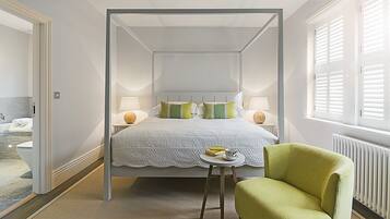 Una cama con dosel, con una colcha y almohadas blancas, una mesita redonda con una taza y libros, un sillón verde y una ventana con persianas.
