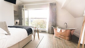 Un dormitorio moderno con una cama grande, una mesita de noche pequeña, una ventana con cortinas y una bañera de cobre.