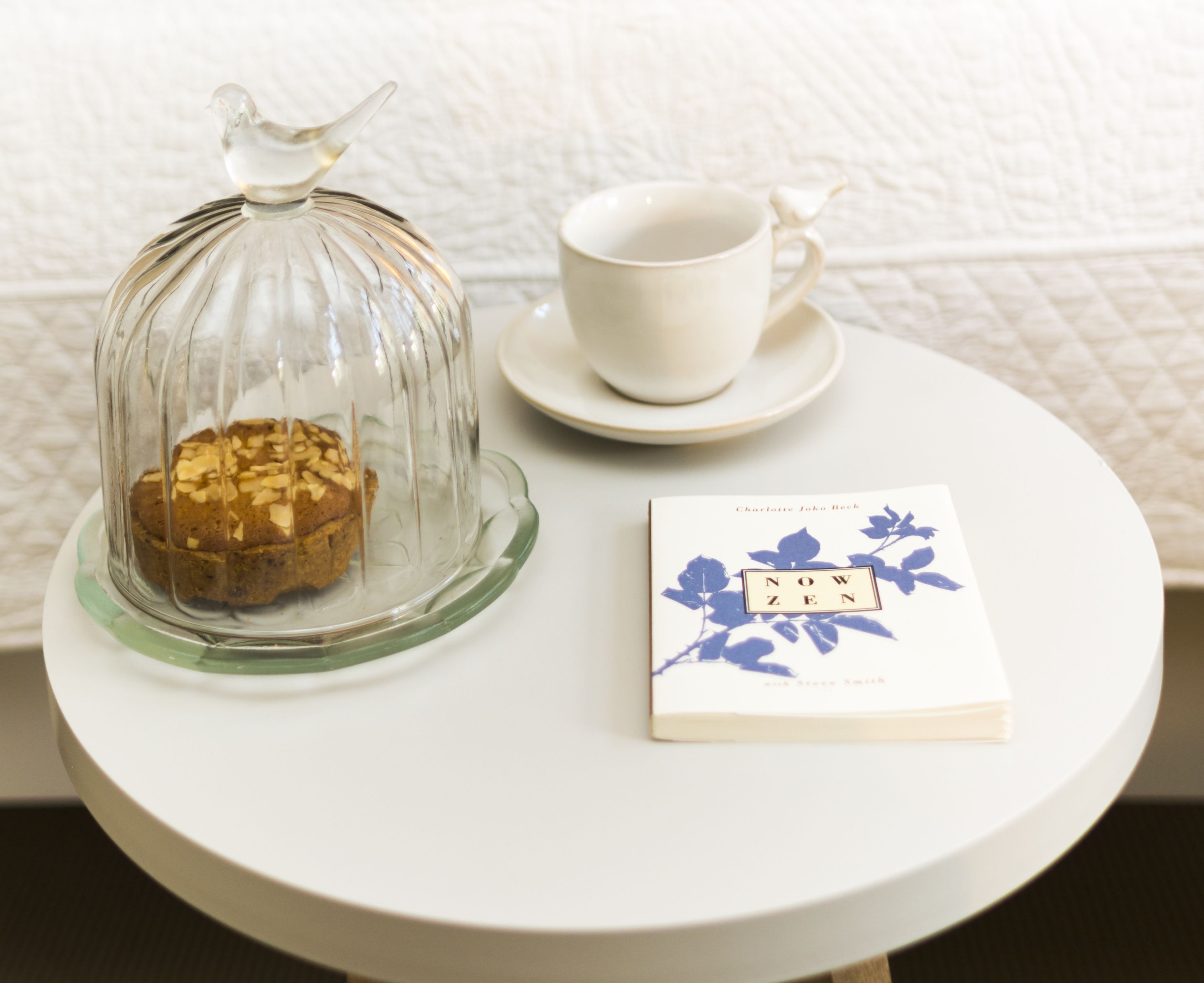 Una mesa redonda con una campana de cristal que contiene una galleta, una taza blanca y un platillo, y un libro titulado 'NOW ZEN' de Charlotte Joka Beck.