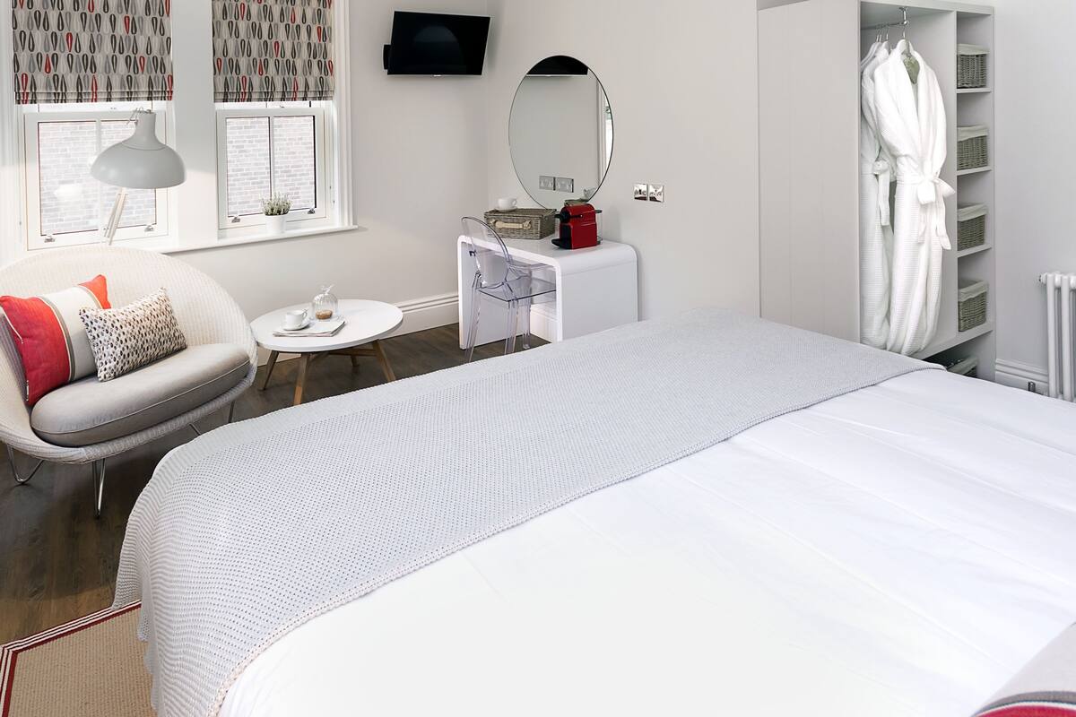 Quarto moderno com uma cama grande, uma cadeira, uma mesinha, um penteoudeiro, um espelho e uma janela com cortinas.