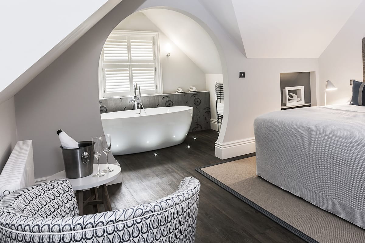 Quarto moderno com cama grande, banheira integrada e área de estar com uma poltrona estampada.