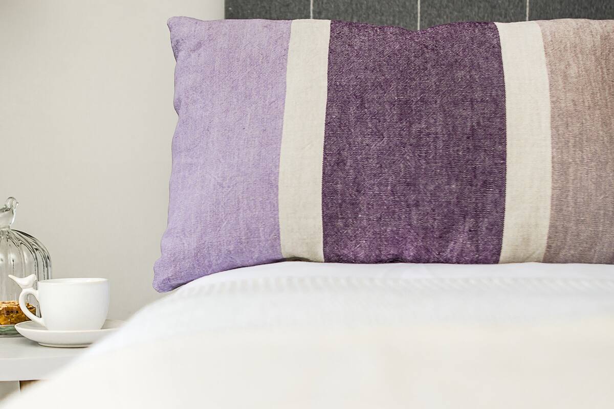 Uma cama bem arrumada com um edredom branco, uma mesa de cabeceira com uma xícara branca e um recipiente de vidro, e uma parede com um travesseiro listrado.