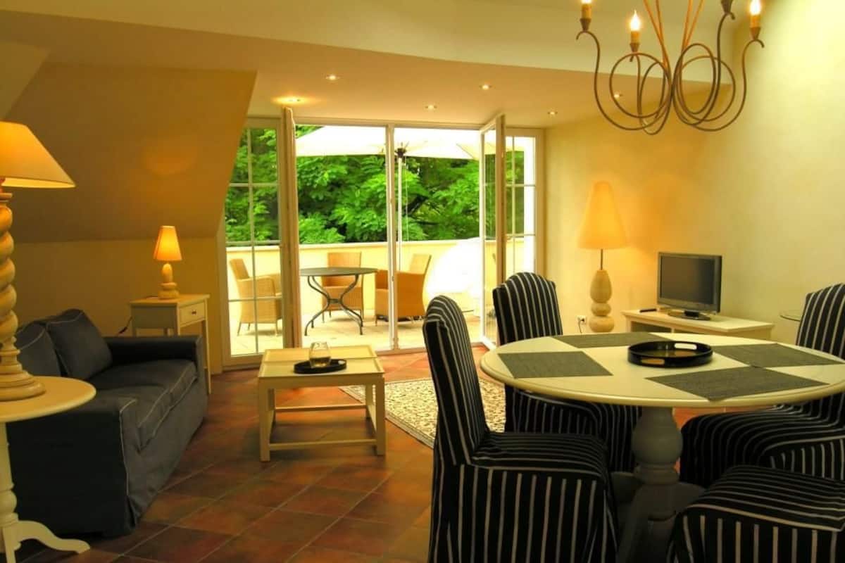 Un salon comprenant un canapé, une table à manger avec des chaises, un téléviseur et un lustre. Une porte vitrée donne accès à un espace extérieur aménagé avec des chaises et une table.