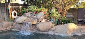A rock waterfall feature in a garden with a wooden fence, potted plants, and outdoor furniture.