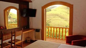 Una stanza con una scrivania, una sedia, una televisione a muro e un balcone con vista su un paesaggio collinare.