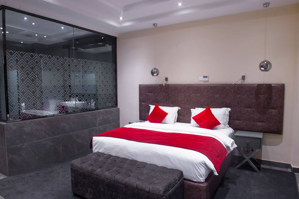 A modern bedroom with a large bed, a grey upholstered bench, a marble-patterned wall, and a glass partition.