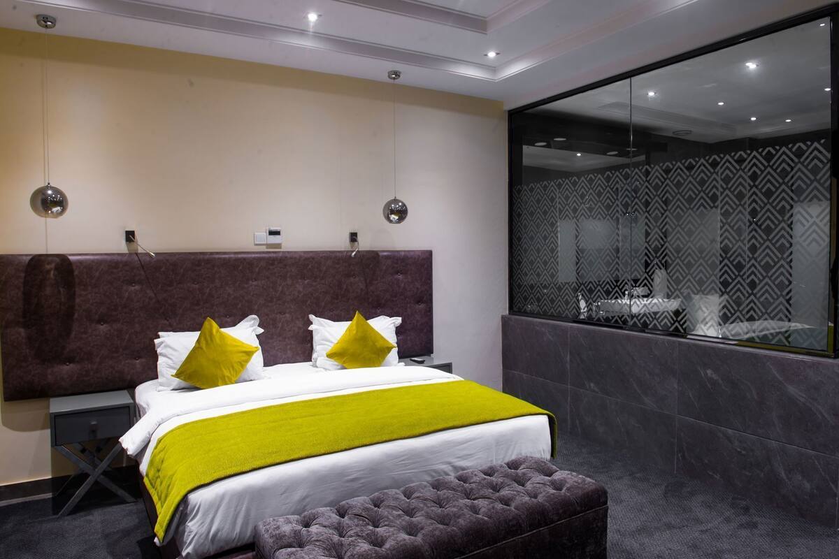 A modern hotel room with a large bed, a grey upholstered bench, a glass shower enclosure, and a patterned wall.