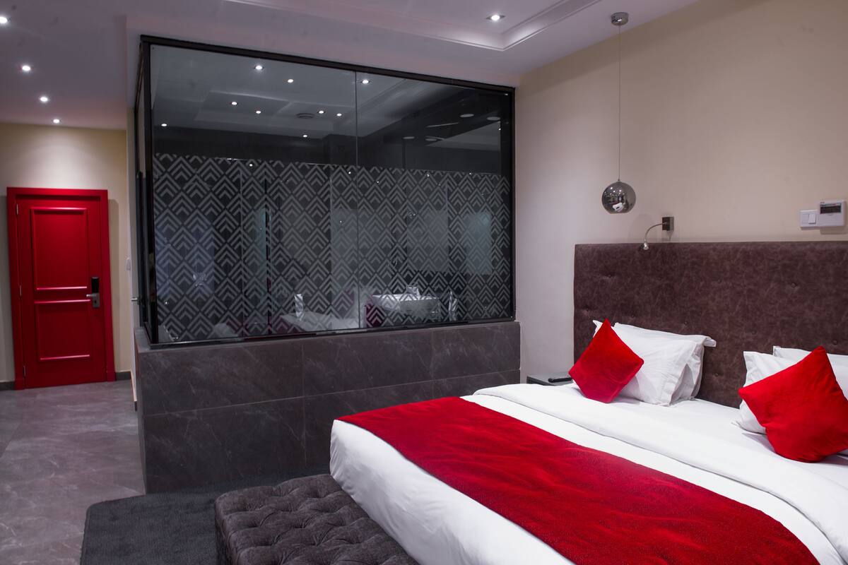 A modern hotel room with a large bed, a red door, and a large window with a patterned view.