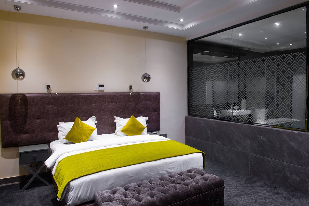 A modern hotel room with a large bed, a bench, a nightstand, and a large mirror.