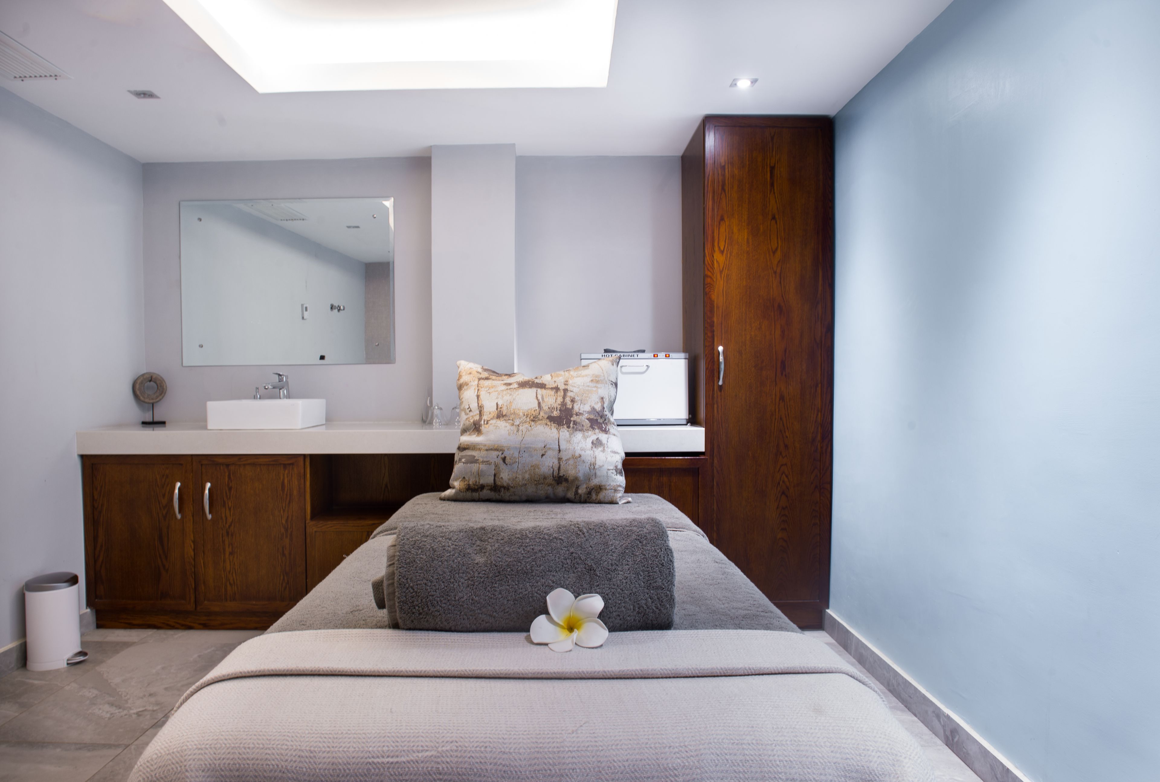A modern bathroom with a large mirror, wooden cabinets, a white sink, and a massage table with a towel and flower.
