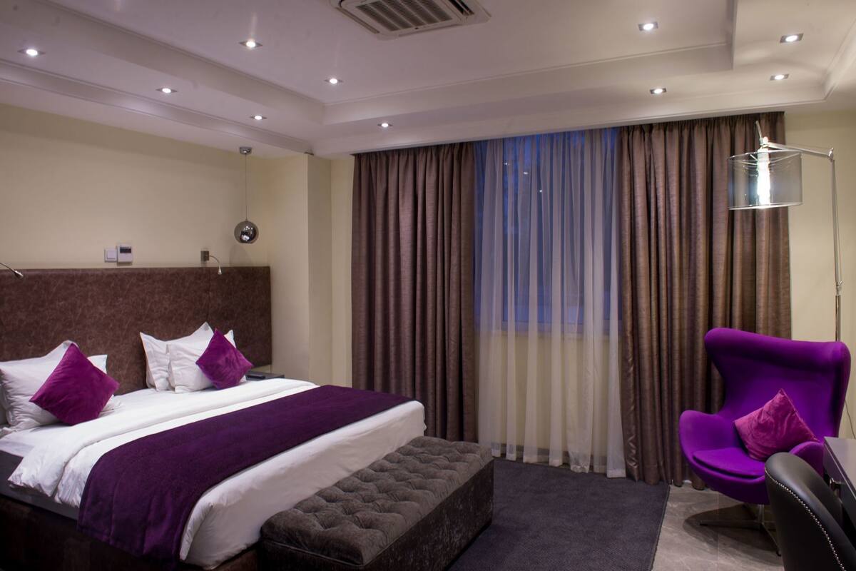 A hotel room with a large bed, a tufted bench, a purple armchair, a desk, and a large window with curtains.