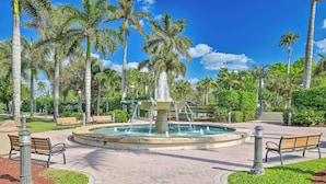 Une fontaine circulaire avec des jets d’eau au centre, entourée de palmiers et de bancs.