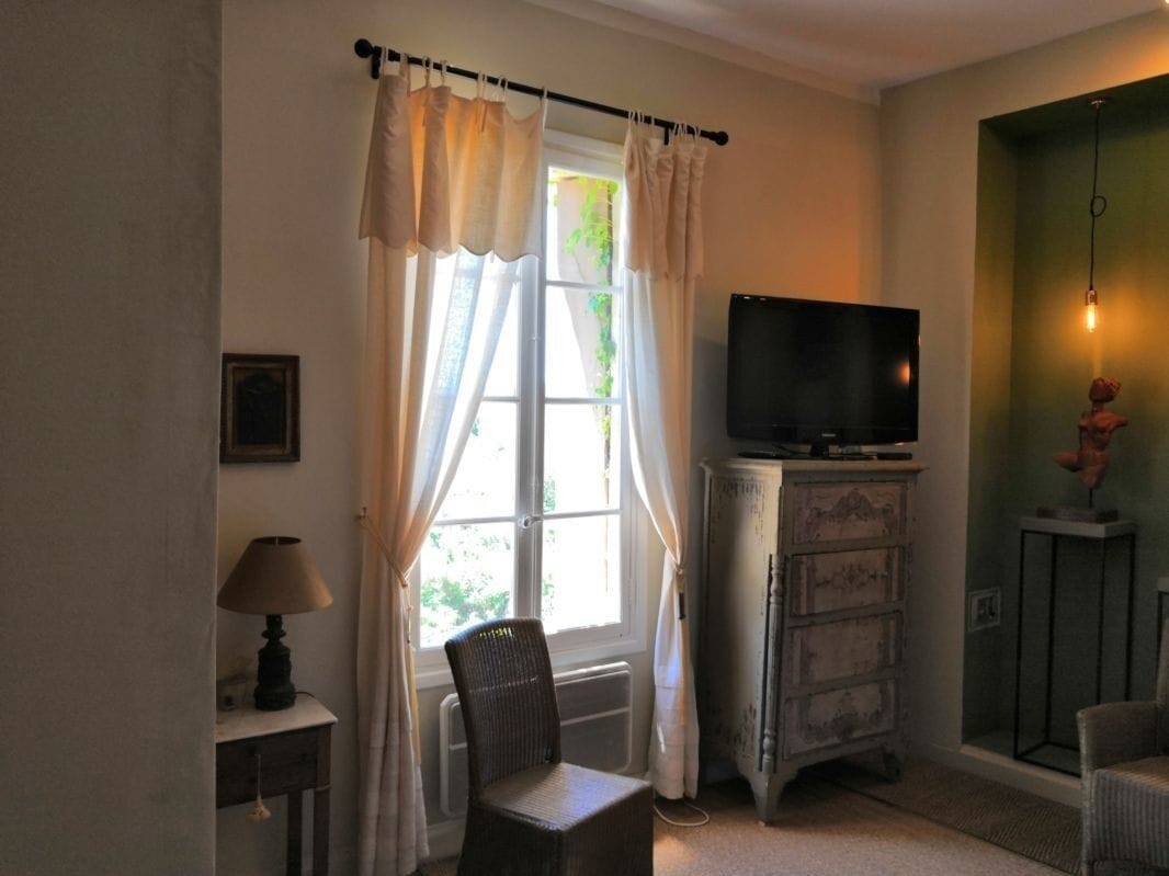 Une pièce avec une télévision posée sur une commode, une chaise, une lampe et une fenêtre avec des rideaux.