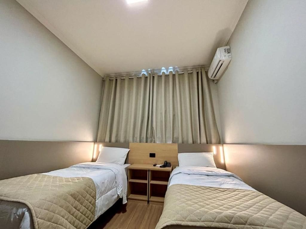 A hotel room with two beds, a bedside table, an air conditioning unit, and a curtain.