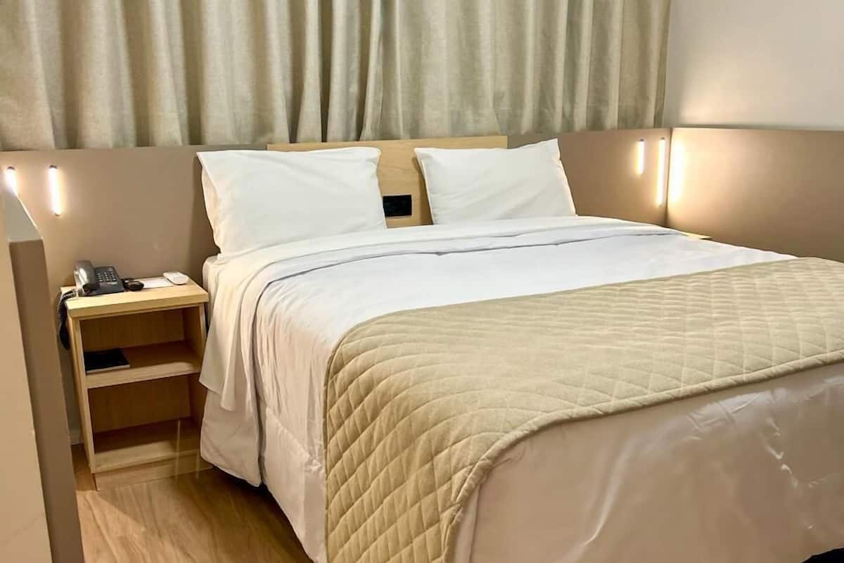 A neatly made bed with a quilted blanket, a bedside table with a lamp, and a curtain covering the window.