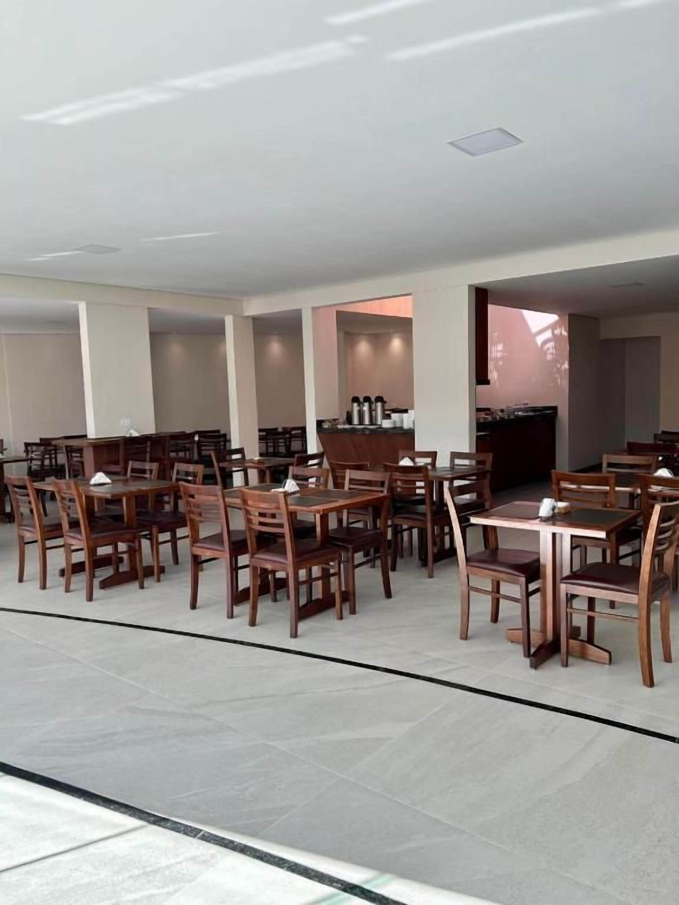 Uma ampla área de refeições interna, com várias mesas e cadeiras de madeira, uma cozinha visível ao fundo e uma vista clara do céu pelas janelas.