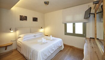 Una cama bien tendida con sábanas blancas, una mesita de noche, un televisor montado en la pared y una ventana con persianas.