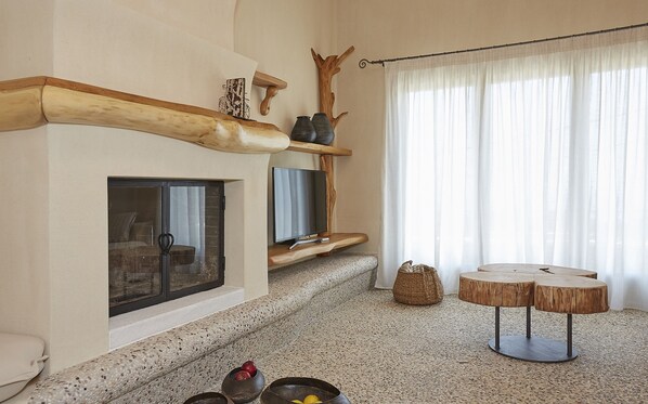 Un salón acogedor con chimenea, televisor, una mesa redonda de madera y un amplio ventanal con cortinas ligeras.